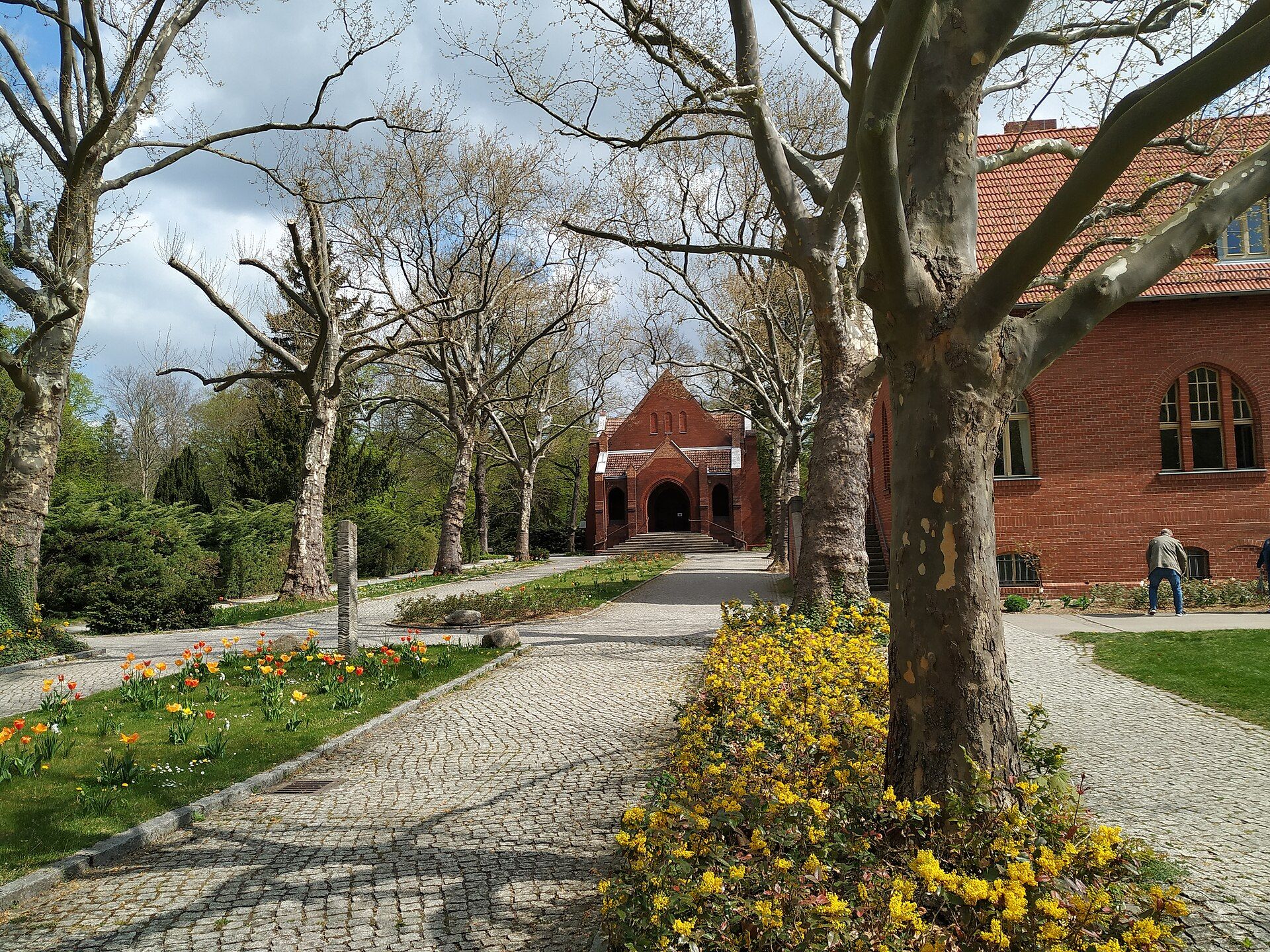 Hauptweg mit Blumenbeeten und Backsteingebäuden auf dem Friedhof Pankow III